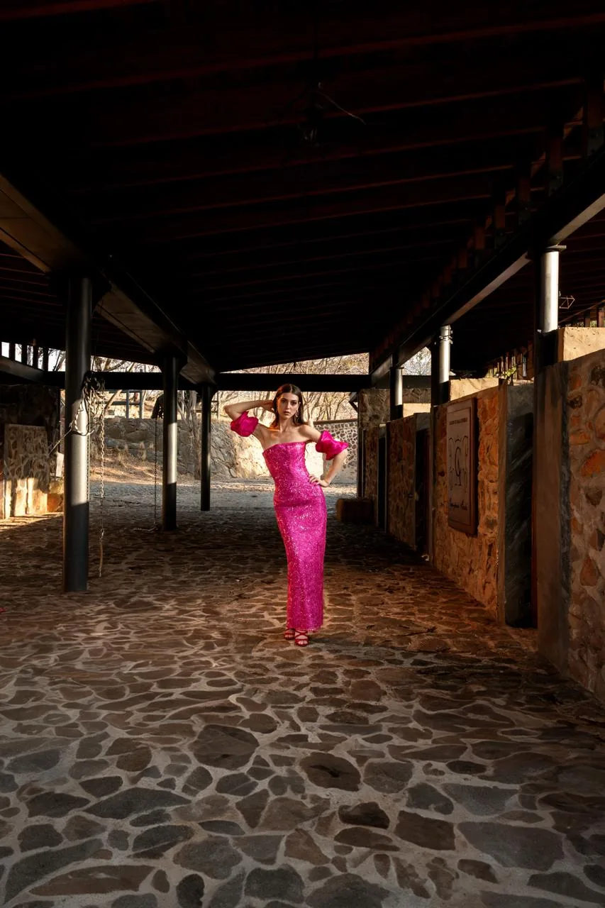 Modelo con vestido fucsia de lentejuelas al fondo del pasillo de caballerizas con piso de piedra