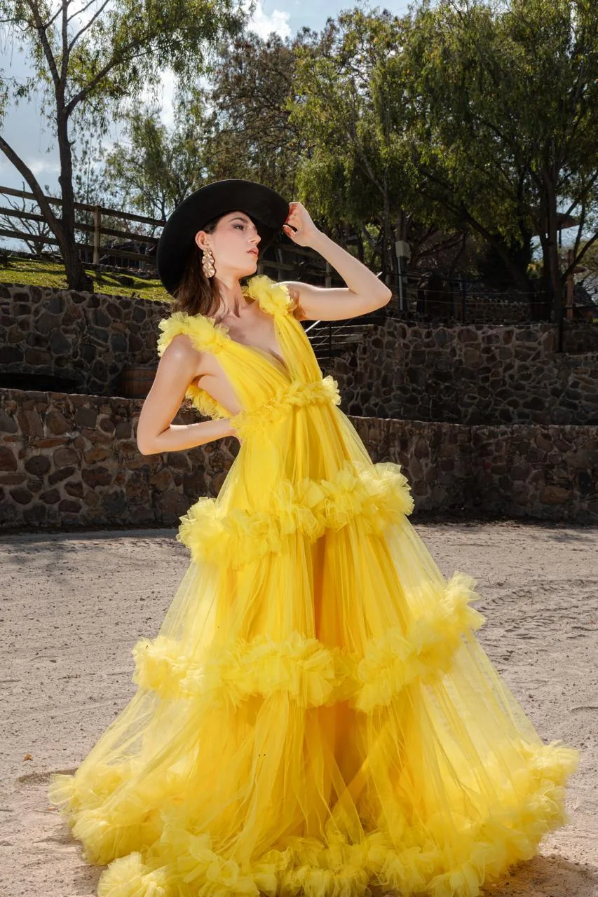 Modelo con vestido amarillo y sombrero negro en la pista, frente al muro de piedra y los árboles